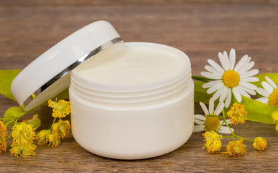 An open white jar of cream with its lid partially off, placed on a wooden surface. The jar is surrounded by vibrant yellow flowers and white daisies with green leaves, emphasizing its natural aroma.