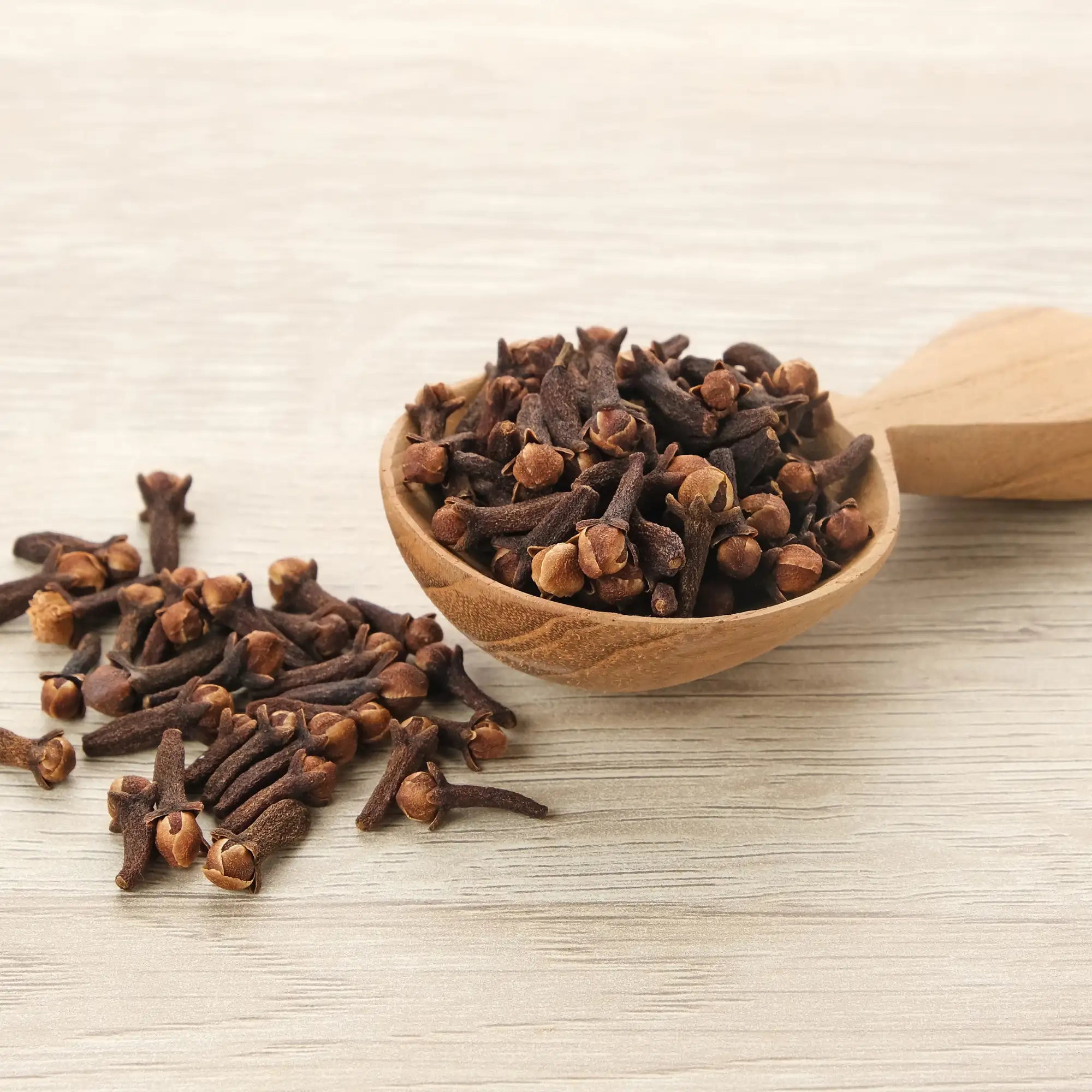 A wooden spoon filled with dried cloves, with additional cloves scattered on a light wooden surface. These aromatic spice buds are the primary source for extracting Clove Oil Essential Oil