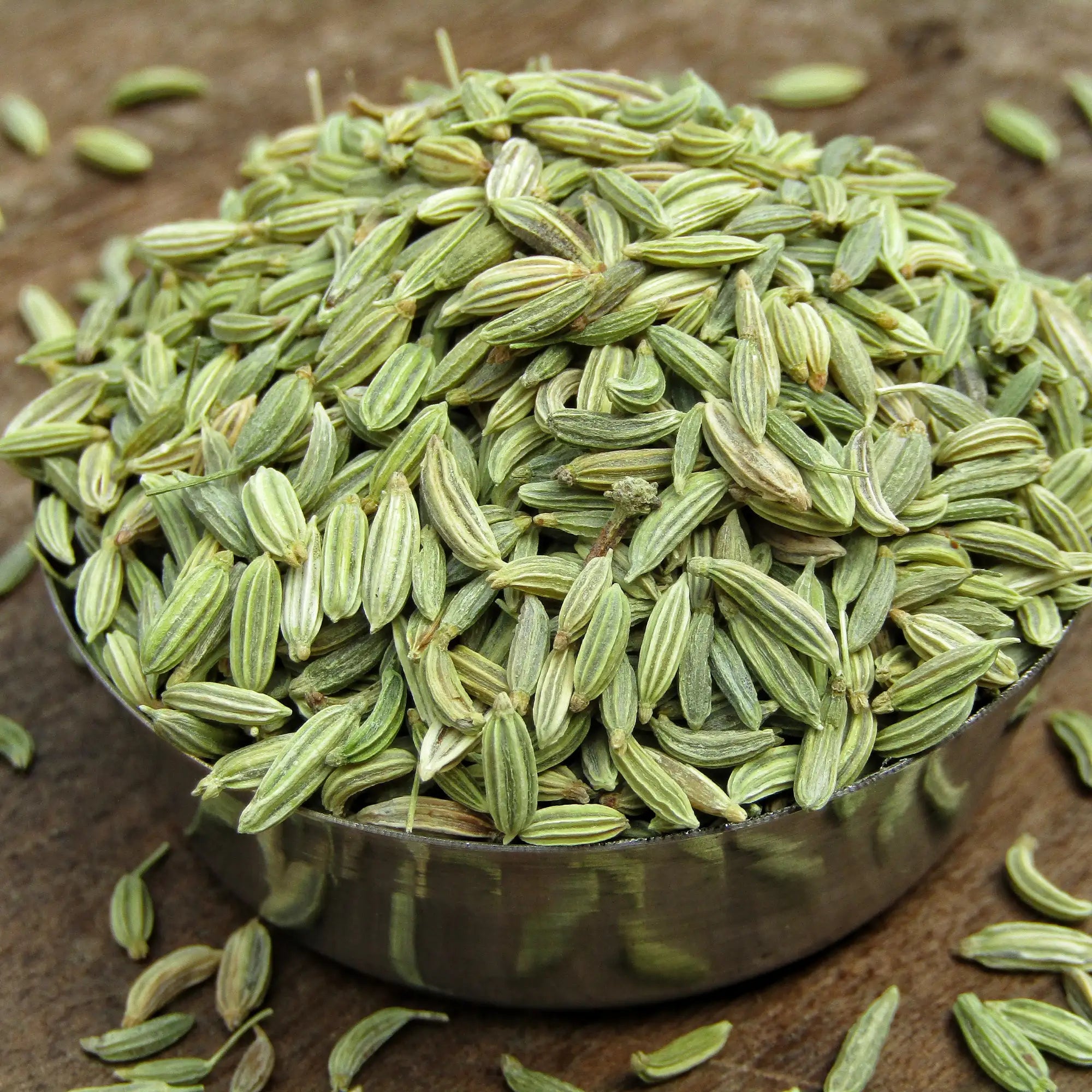A silver bowl overflowingly filled with fennel seeds with additional fennel scattered on a dark wooden surface, that is used by Hiya India to manufacture fennel essential oil.